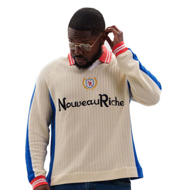 A man wearing a beige ribbed knit sweater with blue and red side panels, blue jeans, and black sneakers, standing against a solid white background.
